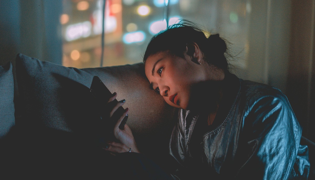 Iemand leunt in het donker met het hoofd tegen een kussen, en kijkt wat verdrietig naar het scherm van de mobiele telefoon.