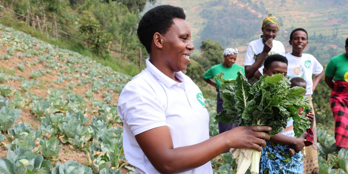 Een vrouw houdt groenten met grote bladeren in haar hand vast, met op de achtergrond andere mensen en daarnaast een veld in de bergen vol met groenten.