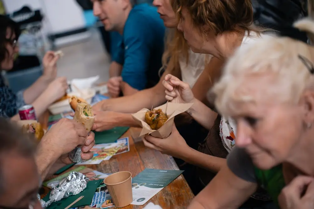 Foto van mensen die aan een tafel met verschillende broodjes in hun handen zitten te eten en praten.