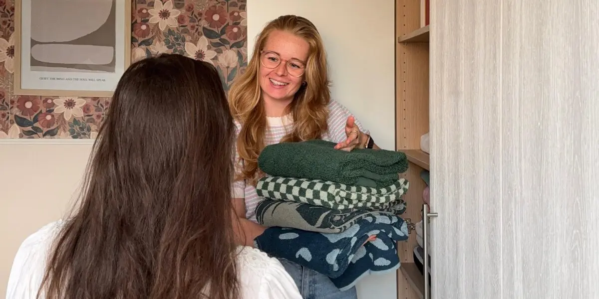 Foto van Saskia van Amerongen staat al pratende met iemand voor een kast en houdt een stapel handdoeken vast.