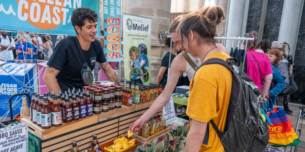 Een persoon presenteert diverse sausjes bij een kraampje. Twee anderen proeven de smaken.