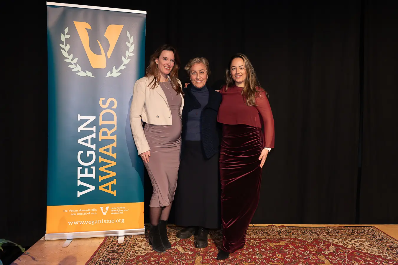 Drie vrouwen poseren lachend voor de Vegan Awards banner.