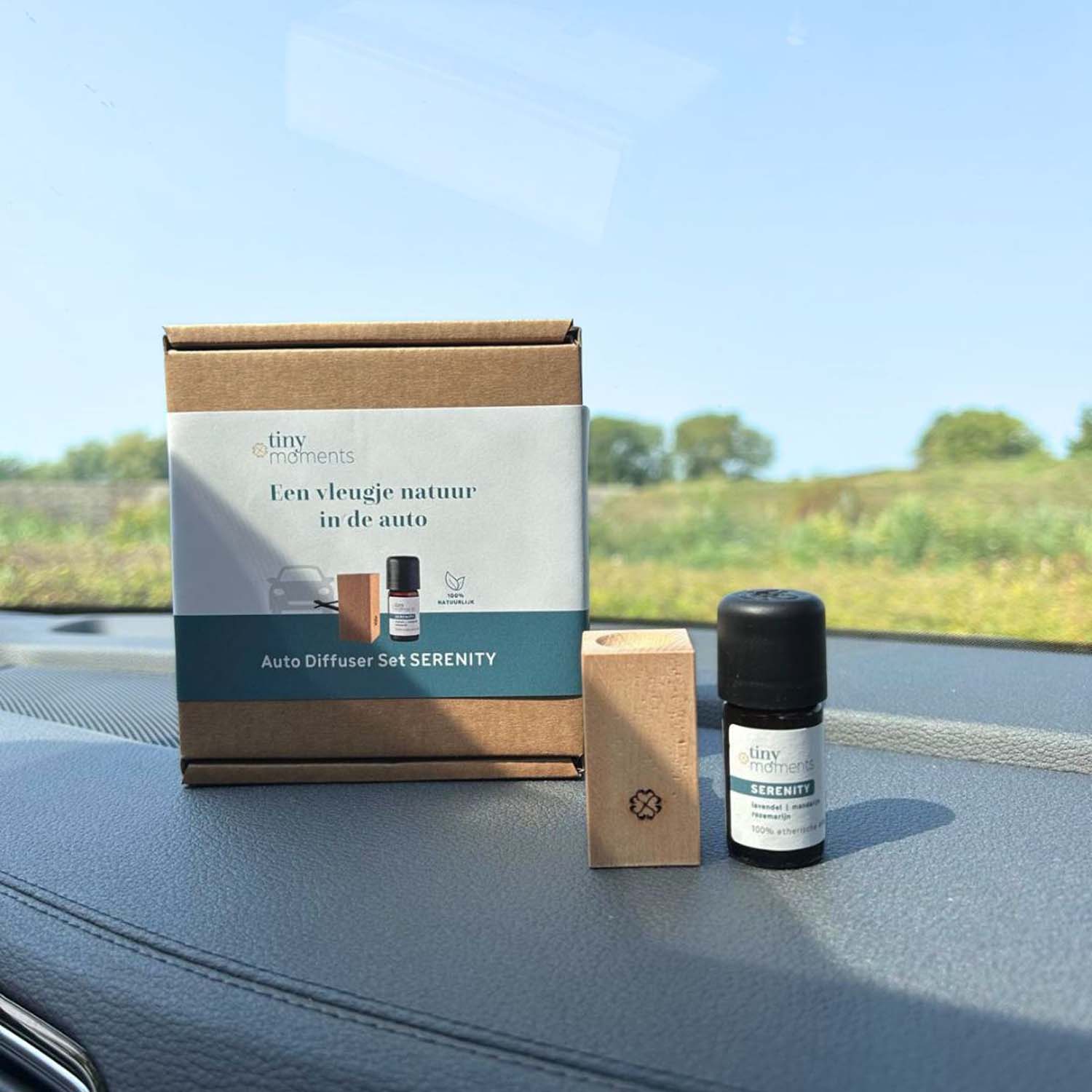 Auto diffuser in verpakking en daarnaast een los flesje en kleine verpakking. Staat op dashboard van een auto. Bomen op de achtergrond