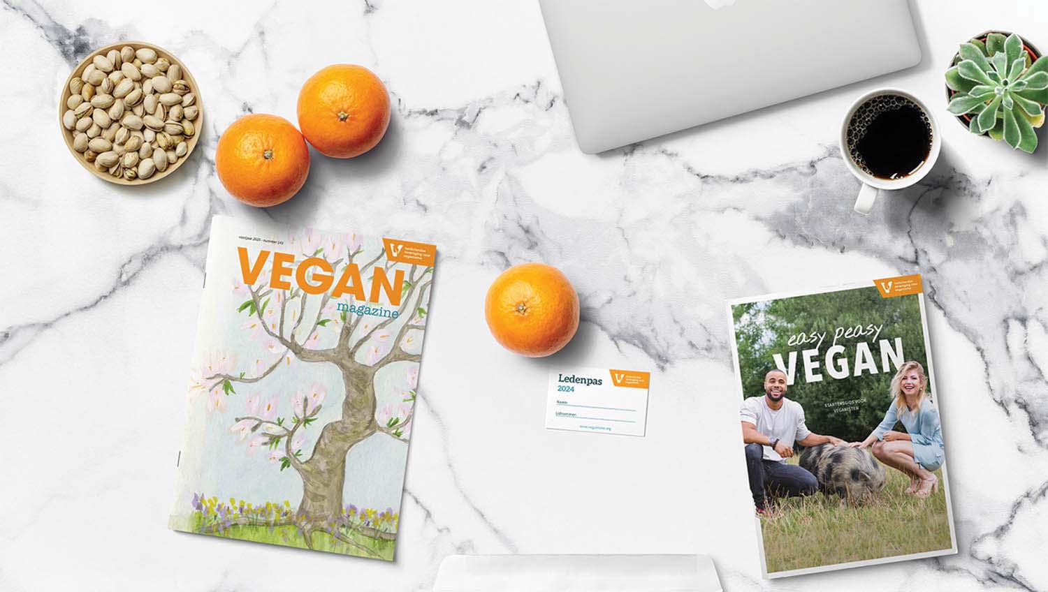 Flatlay van het magazine, Easy peasy vegan startersgids en ledenpas op marmeren tafel, met ook nootjes, plantjes, een laptop en sinaasappels