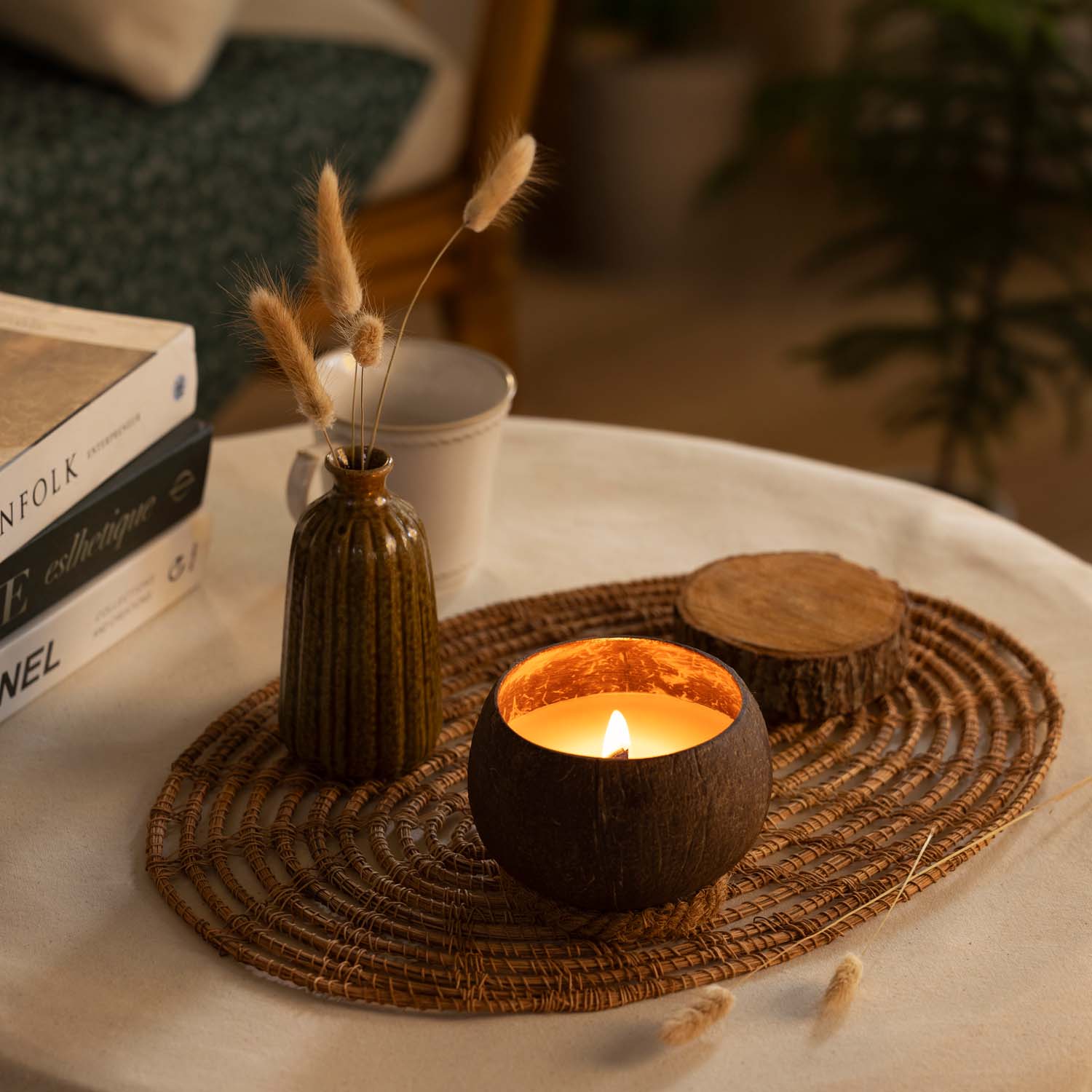 Kokosnoot kaarsje in kokosnoot op een rieten plateau, met ook een vaasje met droogbloemen, houten schijfje, boeken en kopje koffie in beeld, die op ene tafeltje staan